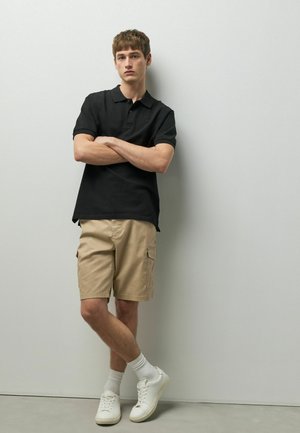 Jeune homme debout contre un mur, portant un polo noir, un short cargo beige, des chaussettes blanches et des baskets blanches, les bras croisés.