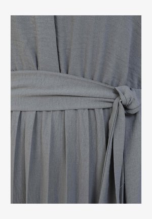 Dettaglio di un vestito in tessuto grigio con texture arricciata, cintura in vita e pieghe sovrapposte. Materiale morbido e leggero con una finitura liscia.