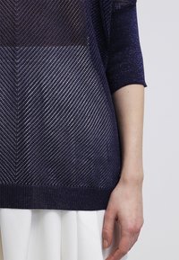 Navy modrý pletený svetr s vzorem chevron, s lesklou texturou a krátkými rukávy. Spodní lem je ribbovaný, s vrstvou bílé látky pod ním.