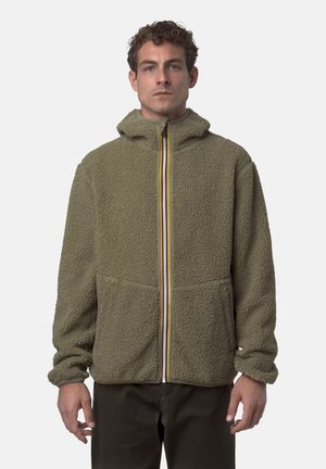 Uomo indossa giacca in pile verde oliva con cappuccio e cerniera a strisce multicolori, in piedi davanti a uno sfondo chiaro e uniforme.