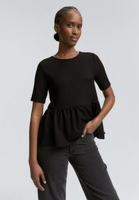 Haut peplum noir à manches courtes, en tissu texturé, avec un col rond et un bas froncé ; associé à un jean gris foncé.