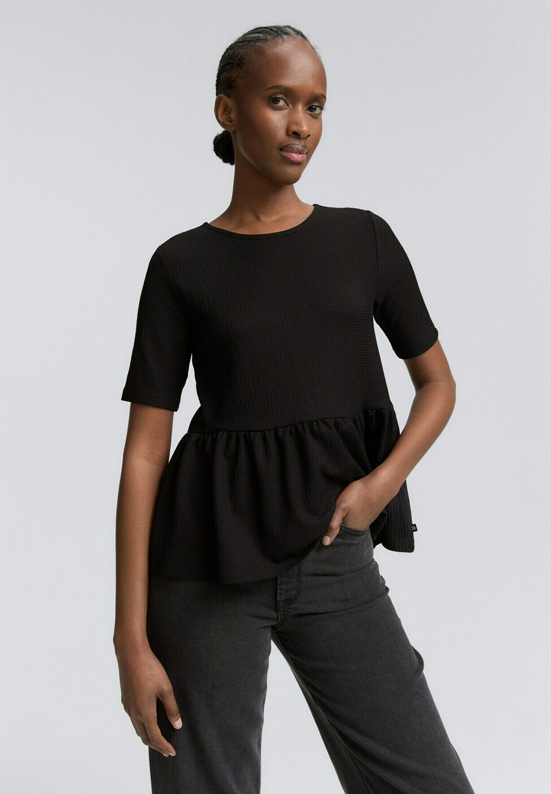 Haut peplum noir à manches courtes, en tissu texturé, avec un col rond et un bas froncé ; associé à un jean gris foncé.