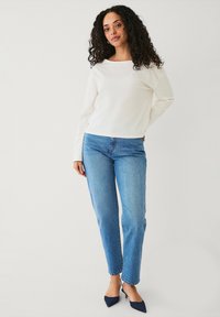 Witte langemouwen top met een bootneck, gecombineerd met blauwe rechte jeans en puntige marineblauwe hakken, gepresenteerd op een model tegen een effen achtergrond.