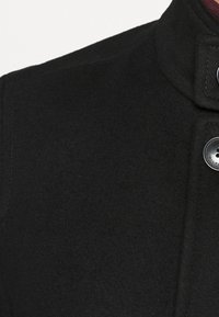 Cappotto nero in misto lana con colletto alto, dotato di chiusura a bottoni e dettagli di cucitura visibili; texture liscia con un design strutturato.