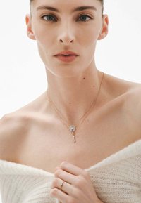 Collar de oro laminado con un colgante rectangular que tiene una piedra clara y un acento colgante. La modelo lleva un suéter blanco con los hombros al descubierto.