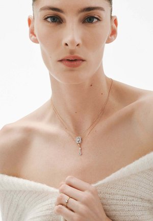 Collar de oro laminado con un colgante rectangular que tiene una piedra clara y un acento colgante. La modelo lleva un suéter blanco con los hombros al descubierto.