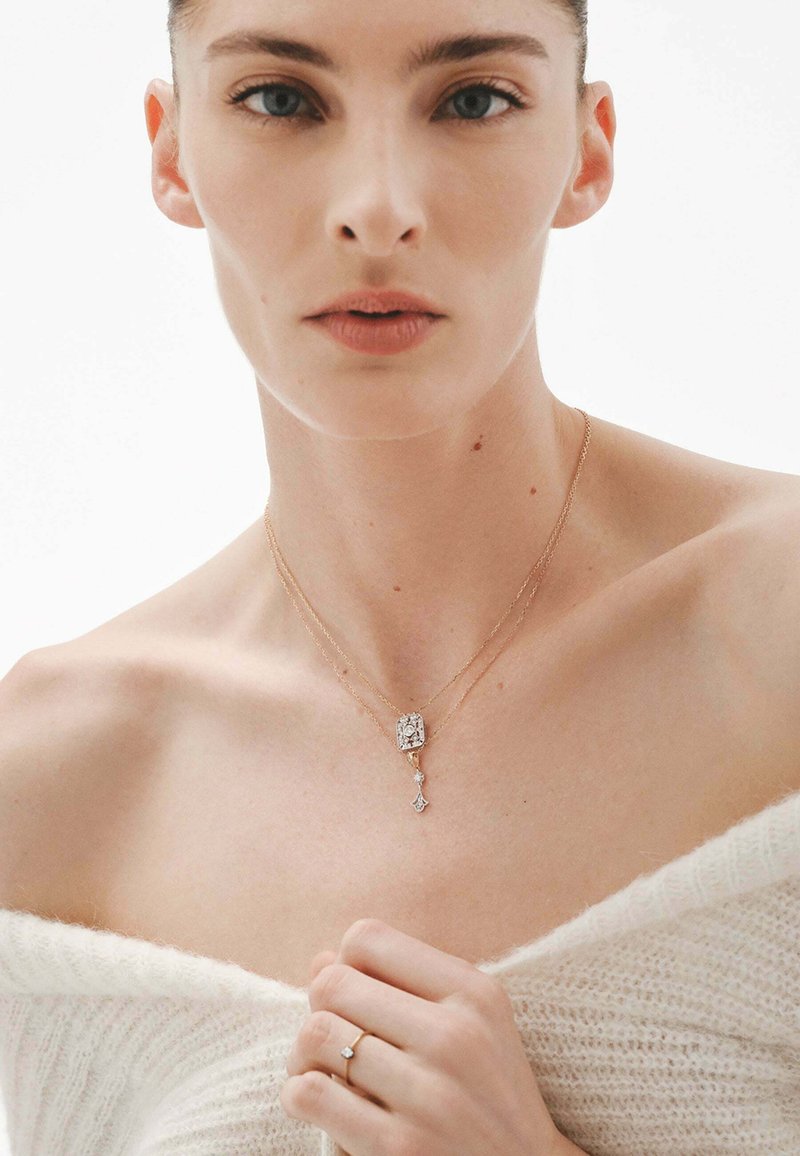 Collar de oro laminado con un colgante rectangular que tiene una piedra clara y un acento colgante. La modelo lleva un suéter blanco con los hombros al descubierto.