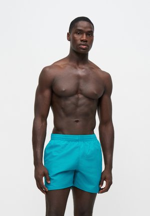 Uomo muscoloso in piedi, indossa pantaloncini da bagno blu brillante, su sfondo bianco uniforme, guarda in avanti con espressione neutra.