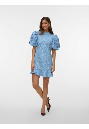 Kvinde iført en blå mini-kjole med blomsterprint, puffede ærmer og flæset kant, kombineret med mørke spidse hæle, stående og smilende.