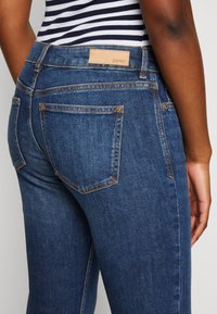 Denimjeans med mörkblå tvätt, med orange sömmar, bakfickor och en tanfärgad läderetikett vid midjan.