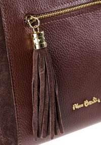 Ingrandimento di una borsa in pelle marrone con dettagli in oro, come la cerniera e il pompon, che mostrano la superficie strutturata e una parte del testo del logo in oro.