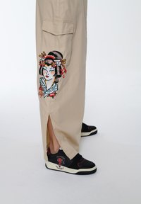 Beige bukser med en broderet geisha-design på venstre side; slidskant, parret med sorte sneaks med røde accenter.