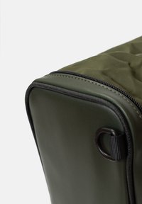 Sac rectangulaire vert avec une surface texturée, doté d'une fermeture éclair noire et d'un bord noir ; comprend une boucle de fixation sur le côté pour le transport.