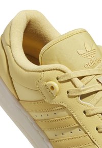 Zapatilla amarilla de Adidas con cuello acolchado, cordones, tres franjas y logo de Adidas en relieve en la lengüeta, mostrada en primer plano.