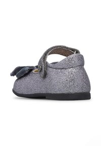 Scarpa argento glitterata con punta arrotondata, caratterizzata da un dettaglio a fiocco e una strap regolabile. Materiale morbido con suola in gomma piatta.