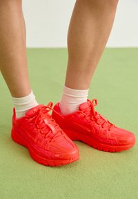 Ryškiai raudoni Nike sportbačiai su tinklo ir gumos detalėmis, su sustiprintais akių užsegimais ir storesniais padais. Avimi su grietinėlės spalvos kojinėmis.
