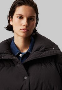 Zwarte pufferjack met een hoge kraag, voorzien van matte knopen en een gladde textuur. Draag hem over een denim overhemd.
