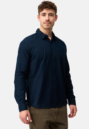 Young man wearing a navy blue buttoned long-sleeve shirt and brown pants, standing against a plain white background.
