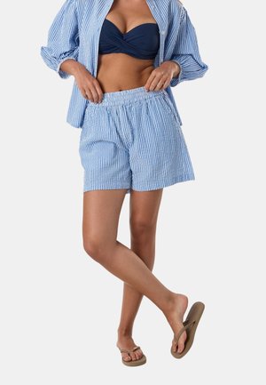 Kvinde iført blå-hvide stribede shorts og matchende åben skjorte, navy bikini top og beige klipklappere, stående med den ene fod løftet.