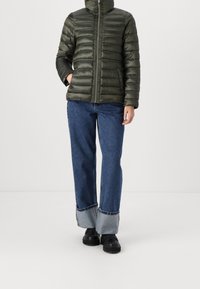 Giacca a vento verde scuro, con colletto alto, motivo a righe orizzontali e zip frontale. Abbinata a jeans blu e scarpe nere.