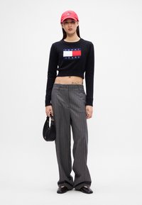 Černý zkrácený svetr s logem "TOMMY JEANS", šedé široké kalhoty s pruhy, červená čepice a černá kabelka, obuté s černými balerínami.