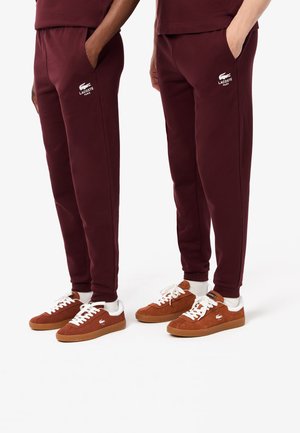 Bordeaux sweatpants van zachte stof, met zijzakken en een wit Lacoste-logo. Gecombineerd met bruine suède sneakers en witte sokken.
