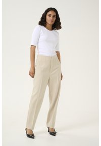 Pantalons beiges ajustés avec des plis à l'avant et une taille haute, associés à un haut ajusté à manches courtes en beige côtelé, et des chaussures noires à talons.