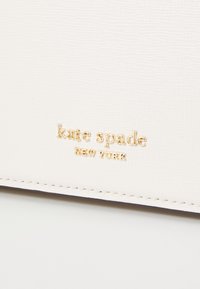 Materiale bianco testurizzato con logo in rilievo "kate spade NEW YORK" in oro. Cuciture visibili lungo il bordo. Design semplice ed elegante.