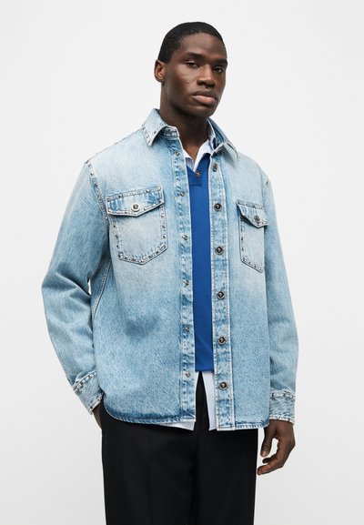 Νέος άνδρας που φοράει μια ανοιχτό μπλε τζιν ζακέτα με καρφιά πάνω από ένα μπλε πουλόβερ και πουκάμισο με ρίγες, στέκεται με το ένα χέρι στην τσέπη.