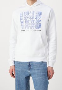 Bílá mikina s kapucí vyrobená z měkké látky s přední kapsou. V přední části se nacházejí modře vytištěné souřadnice a text „NEVER STOP EXPLORING“.