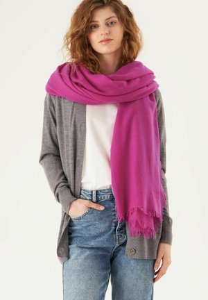 Écharpe magenta avec des bordures frangées posée sur un cardigan gris, porté sur un haut blanc, associé à un jean bleu clair.