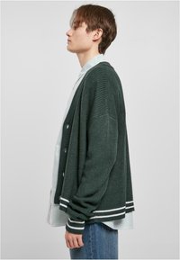 Grønn strikket cardigan med avslappet passform, med hvite stripete detaljer på ermene og knappestolpe foran, lagd oppå en blå stripete skjorte.