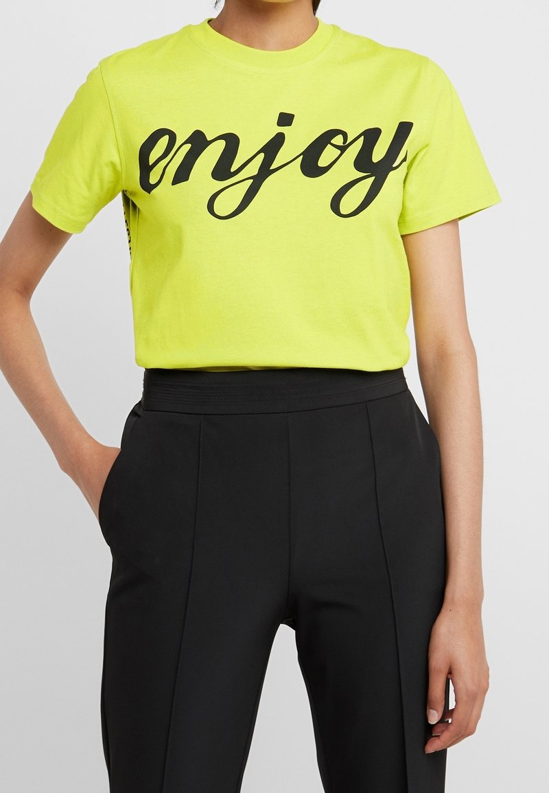 Camiseta de algodón amarilla neón con un texto en cursiva negra que dice "disfruta". Combina con unos pantalones negros de talle alto, que muestran una textura suave.