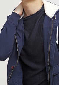 Chaqueta azul marino con cierre de cremallera y forro de forro polar blanco, con capucha, cordones y un botón redondo marrón en la parte delantera. Se lleva sobre una camiseta oscura.