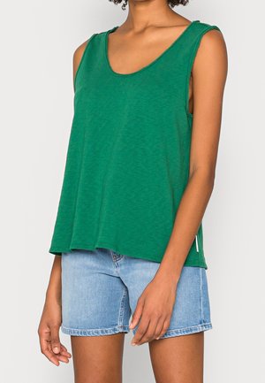 Vrouw draagt een los groen mouwloos topje en lichtblauwe denimshorts, staand tegen een effen lichte achtergrond.
