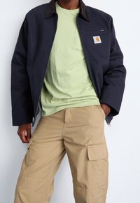 Marineblaue Jacke mit braunem Kragen, Reißverschlusstasche und Logo-Aufnäher, kombiniert mit einem hellgrünen Hemd und beigen Cargo-Hosen.