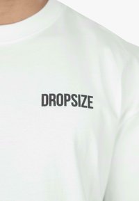 Weißes Baumwoll-T-Shirt mit einem schwarzen, gedruckten "DROPSIZE"-Logo auf der linken Brustseite. Rundhalsausschnitt, weiche Textur, lässige Passform.