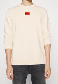 Sudadera beige claro de manga larga, con puños y dobladillo acanalados. Presenta un parche rectangular rojo con la palabra "HUGO" en negro.