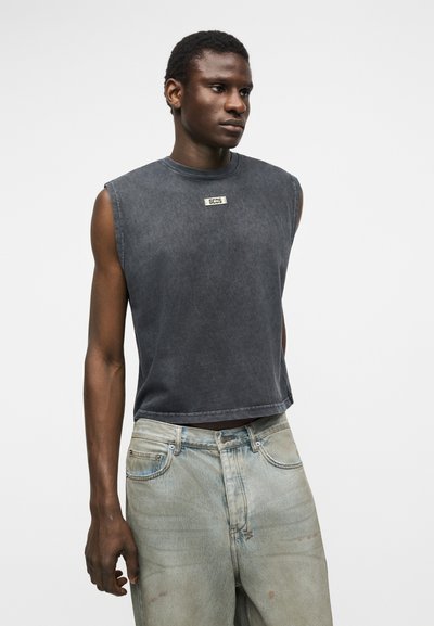 Νέος άνδρας με αμάνικο σκούρο γκρι πουκάμισο με ετικέτα "GCDS" και ανοιχτόχρωμο ξεθωριασμένο τζιν, στέκεται μπροστά σε λευκό απλό φόντο.