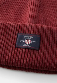 Burgundy ribbed knit beanie dengan cuff yang dilipat, menampilkan label biru navy dengan logo GANT berwarna putih dan merah serta teks "New Haven Connecticut".