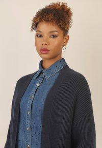 Granatowy, dzianinowy kardigan noszony na dżinsowej koszuli z metalowymi ćwiekami, z kołnierzykiem i zapięciem na guziki, o gładkiej fakturze.