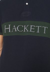 Marinblå pikétröja med ett grönt horisontellt rand. Har ordet "HACKETT" i broderad rutmönster. Tvåknappsdetalj vid kragen.