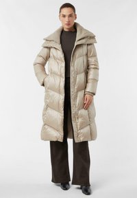 Beige lange pufferjas met een hoge kraag, gewatteerd ontwerp en knopen aan de voorkant. Gecombineerd met donker brede broekspijpen en zwarte schoenen.