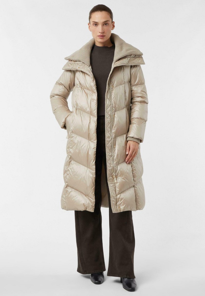 Beige lange pufferjas met een hoge kraag, gewatteerd ontwerp en knopen aan de voorkant. Gecombineerd met donker brede broekspijpen en zwarte schoenen.