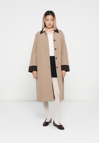 Beiger Trenchcoat mit braunem Kragen, mit geradem Schnitt, großen Knöpfen und weiten Ärmeln. Tragen über einem weißen Hemd und Shorts.