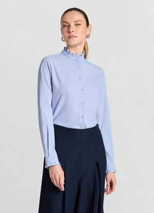 Hellblaue Bluse mit hohem Rüschenkragen und langen Ärmeln, mit Knopfleiste vorne und gerüschten Ärmelbündchen, kombiniert mit einer dunklen Hose.