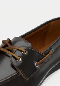 Gros plan d’une chaussure en cuir marron foncé avec des lacets en cuir beige passés dans des œillets métalliques et une doublure intérieure beige lisse.