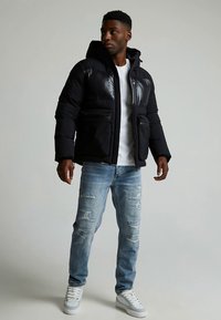 Černá péřová bunda s lesklým panelem, má kapuci, dvě přední kapsy na zip, nošena přes bílou košili a roztrhané modré džíny.