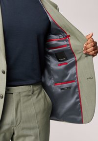 Jasnozielona marynarka w kolorze z fakturowanej tkaniny, szare podszewki, czerwone akcenty i wiele wewnętrznych kieszeni. Pasujące jasnozielone spodnie.