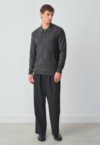 Szürke pulóver, negyed cipzárral, texturált gyémántmintával. Lüktető fekete nadrággal és fekete cipővel viselve.